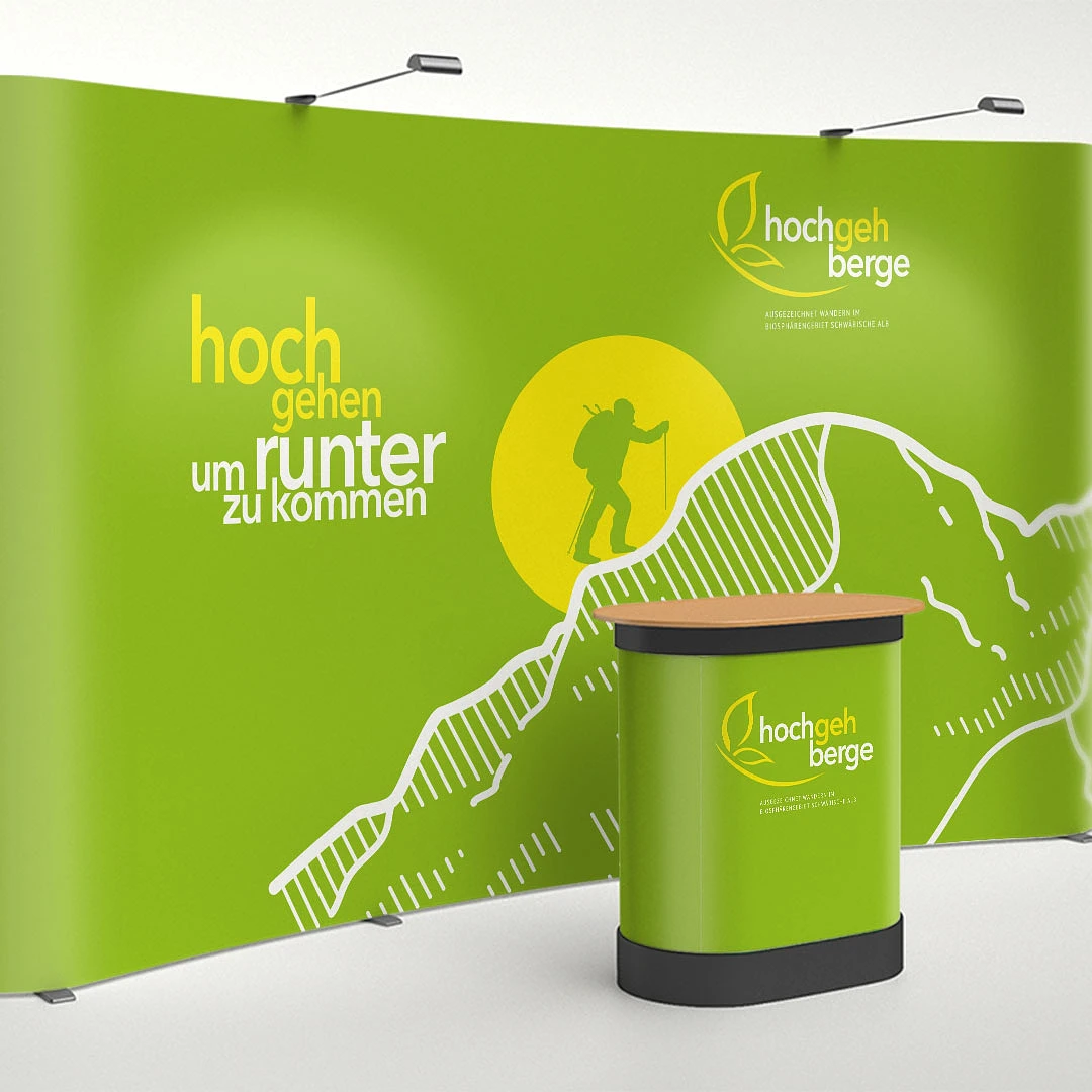 geschwungene grüne messewand mit dem slogan »hochgehen um runterzukommen«, wandererfigur und hügelgrafik, davor ein passender thekenaufsteller